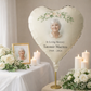 Heart-shaped balloon with a photo of an elderly woman and memorial text, surrounded by flowers and candles on a table.