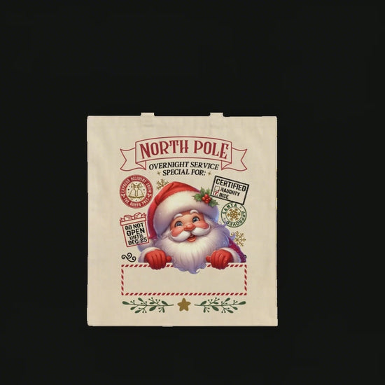 Top-down video of a North Pole Santa tote bag on a wooden table, with a woman’s hands placing small wrapped gifts, a candy cane, and a white coffee cup on it. A child’s hand briefly takes a candy, creating a cozy Christmas family moment.